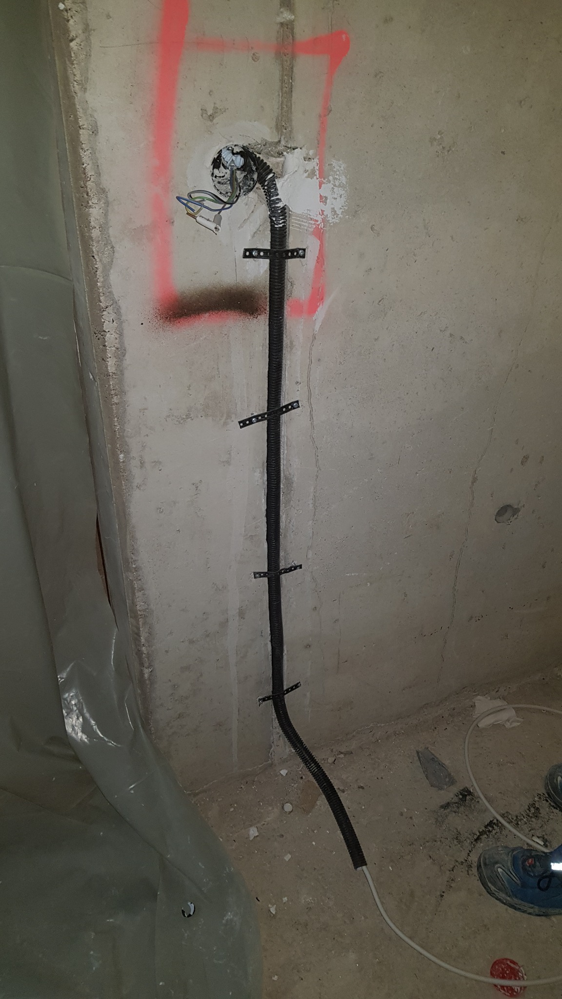 Kabel elektryczny w karbowanej rurze przymocowany do surowej ściany z betonu za pomocą metalowych uchwytów, widoczne wystające przewody i oznaczenia czerwoną farbą.