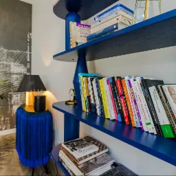 Niebieski regał z książkami, lampka nocna z abażurem i roślina doniczkowa tworzą przytulny kącik w nowoczesnym wnętrzu, z tapetą imitującą szkic miasta w tle.