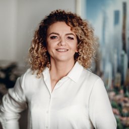 WARSO NIERUCHOMOŚCI - Portret uśmiechniętej kobiety z burzą blond loków, ubrana w białą koszulę, na tle abstrakcyjnego obrazu przedstawiającego panoramę miejską.