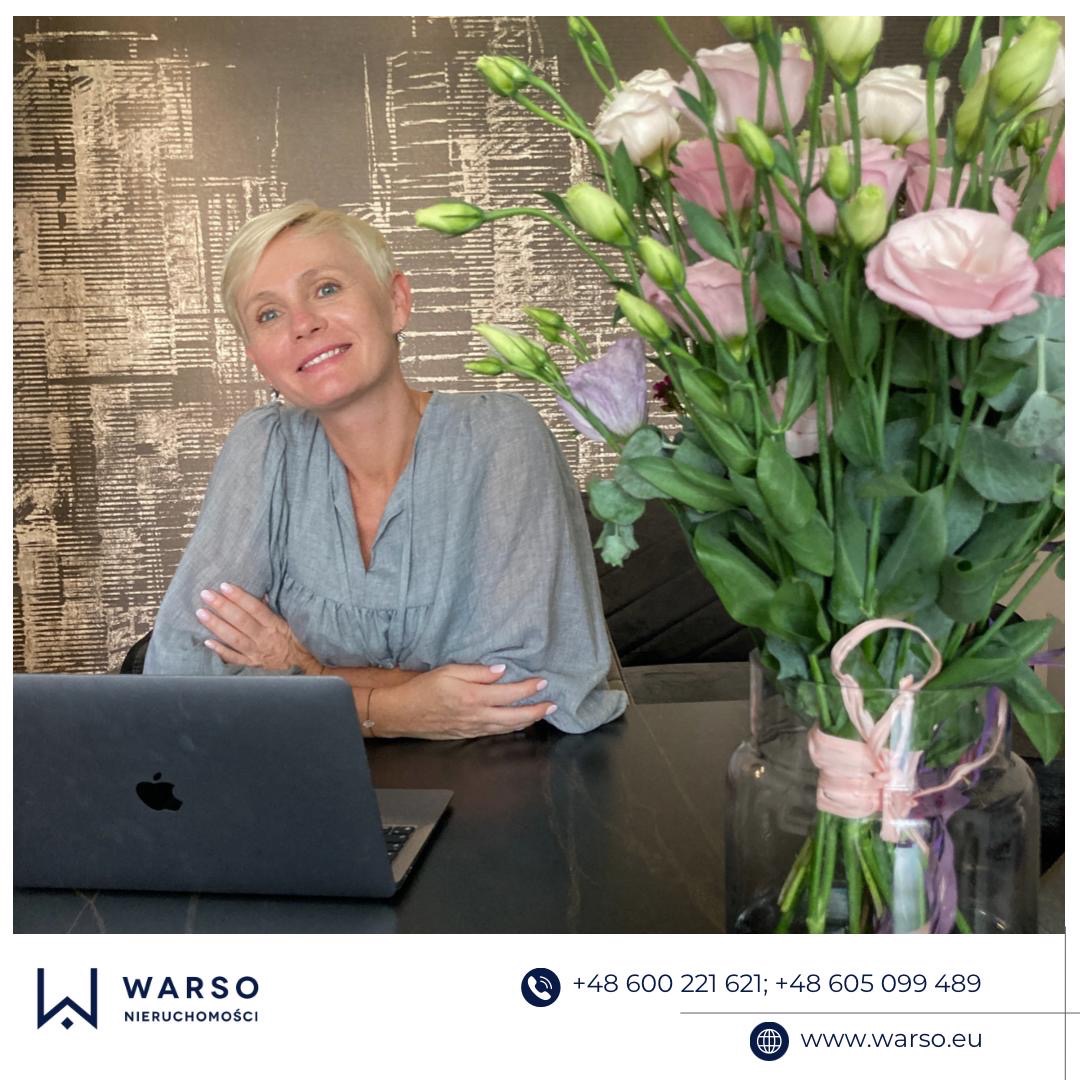 Uśmiechnięta kobieta z krótkimi blond włosami siedzi za biurkiem z laptopem i bukietem różowych kwiatów w wazonie, na tle wzorzystej tapety, prezentująca ofertę agencji nieruchomości.