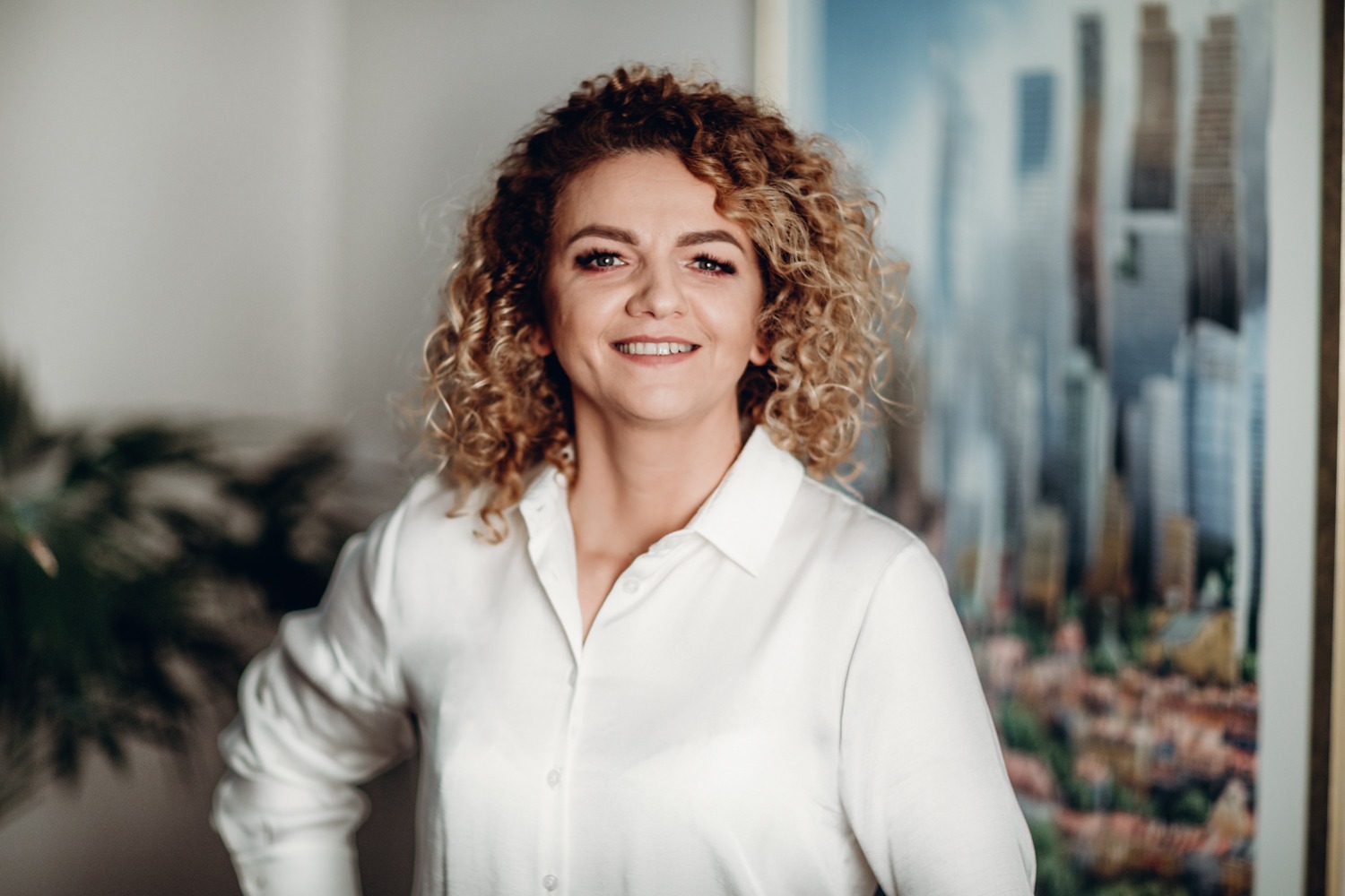 Portret uśmiechniętej kobiety z burzą blond loków, ubrana w białą koszulę, na tle abstrakcyjnego obrazu przedstawiającego panoramę miejską.