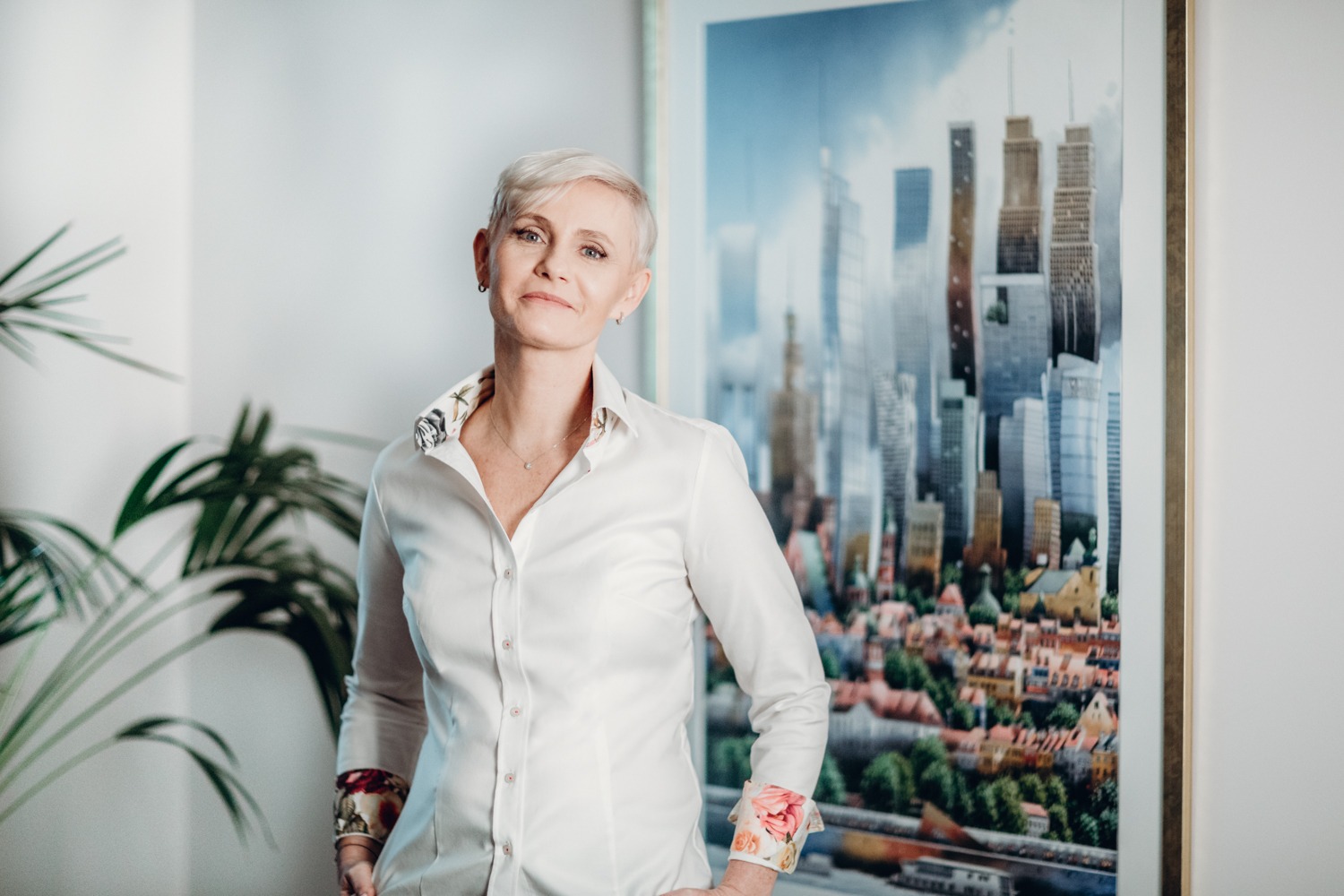 Kobieta z krótkimi siwymi włosami w białej koszuli z kwiatowymi mankietami i kołnierzykiem, stojąca na tle obrazu przedstawiającego panoramę miasta i rośliny doniczkowej.