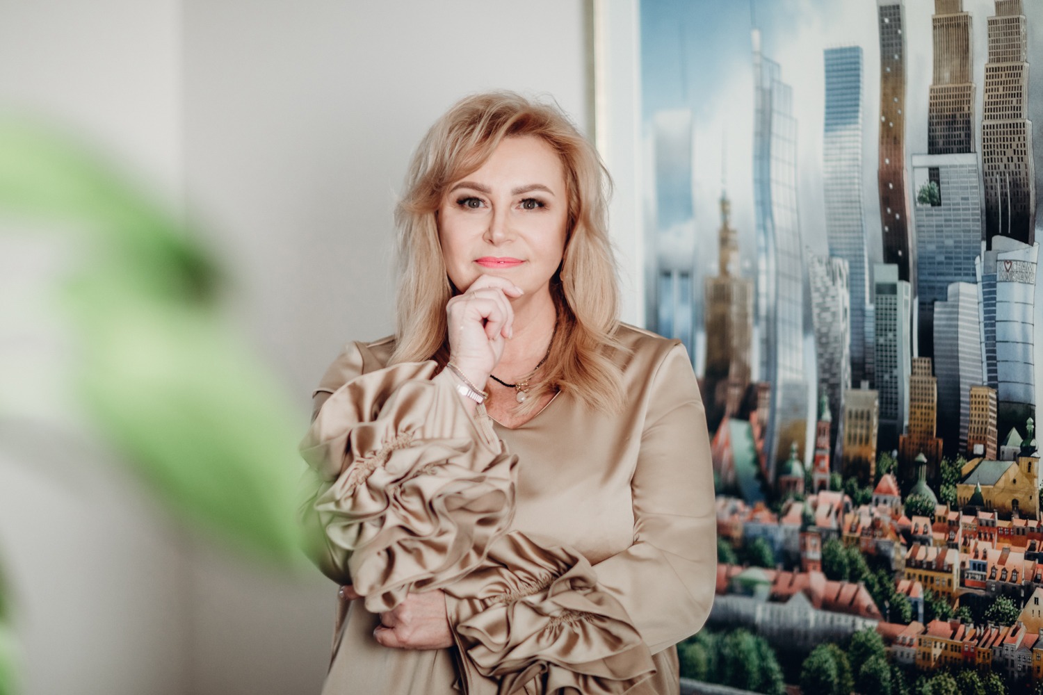 Portret kobiety w beżowej sukience z bufkami na tle obrazu przedstawiającego panoramę miasta z wysokimi budynkami i starymi kamienicami.