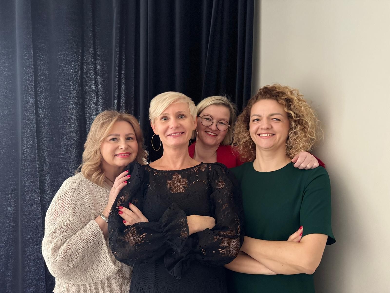 Portret grupowy czterech uśmiechniętych kobiet, w tym jedna w okularach, na tle ciemnej zasłony i jasnej ściany.