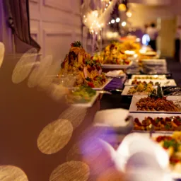 Bogato zastawiony stół szwedzki z różnorodnymi potrawami na białych talerzach, dekoracyjnie oświetlony, z rozmytymi elementami w pierwszym planie.