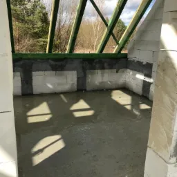 Wnętrze domu w trakcie budowy: ściany z białych bloczków, świeżo wylana betonowa podłoga z widocznymi cieniami, zielona konstrukcja dachu, widok na drzewa w tle.