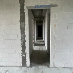 Wnętrze surowego domu w trakcie budowy: ściany z białych bloczków, widoczny fragment zbrojenia i niebieski napis na ścianie nad otworem drzwiowym.