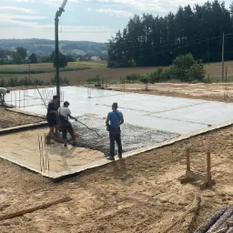 Wylewanie betonu na przygotowany fundament z widocznymi prętami zbrojeniowymi i pracownikami budowlanymi, w tle krajobraz zalesiony.