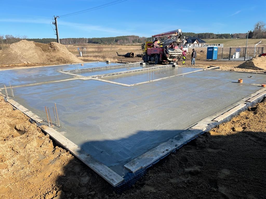 Wylany fundament pod budowę domu jednorodzinnego z widocznym zbrojeniem i pompą do betonu na placu budowy w słoneczny dzień.