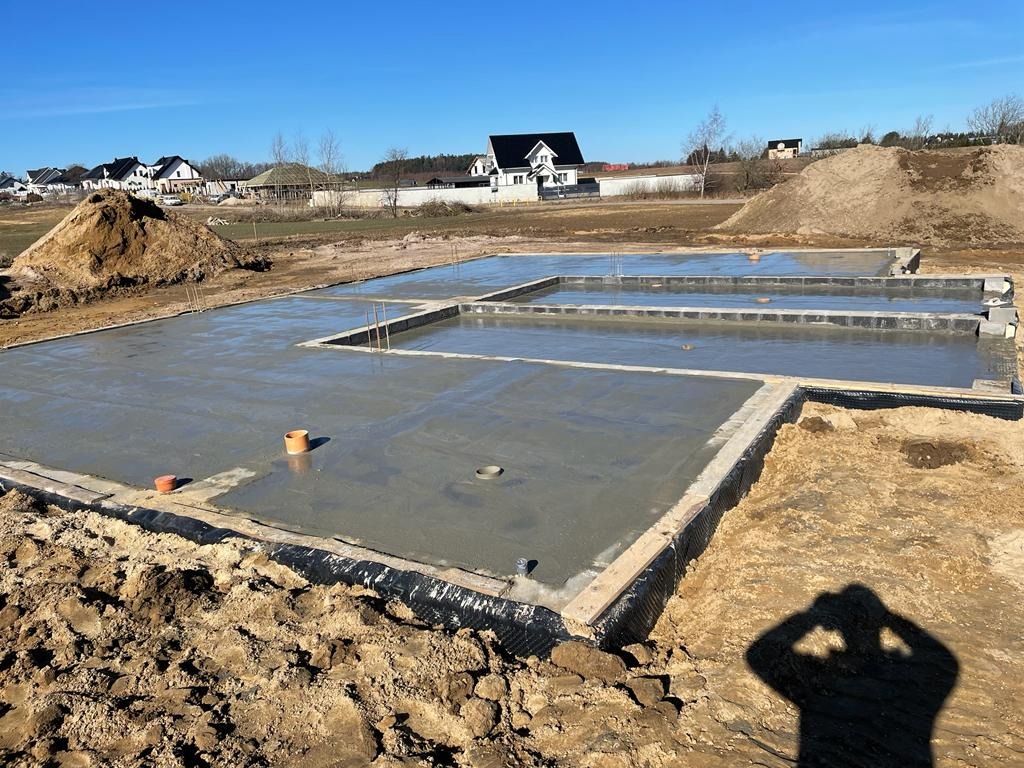 Wylany fundament pod budynek jednorodzinny, widoczne szalunki i wystające pręty zbrojeniowe, w tle zabudowa jednorodzinna i hałda ziemi.