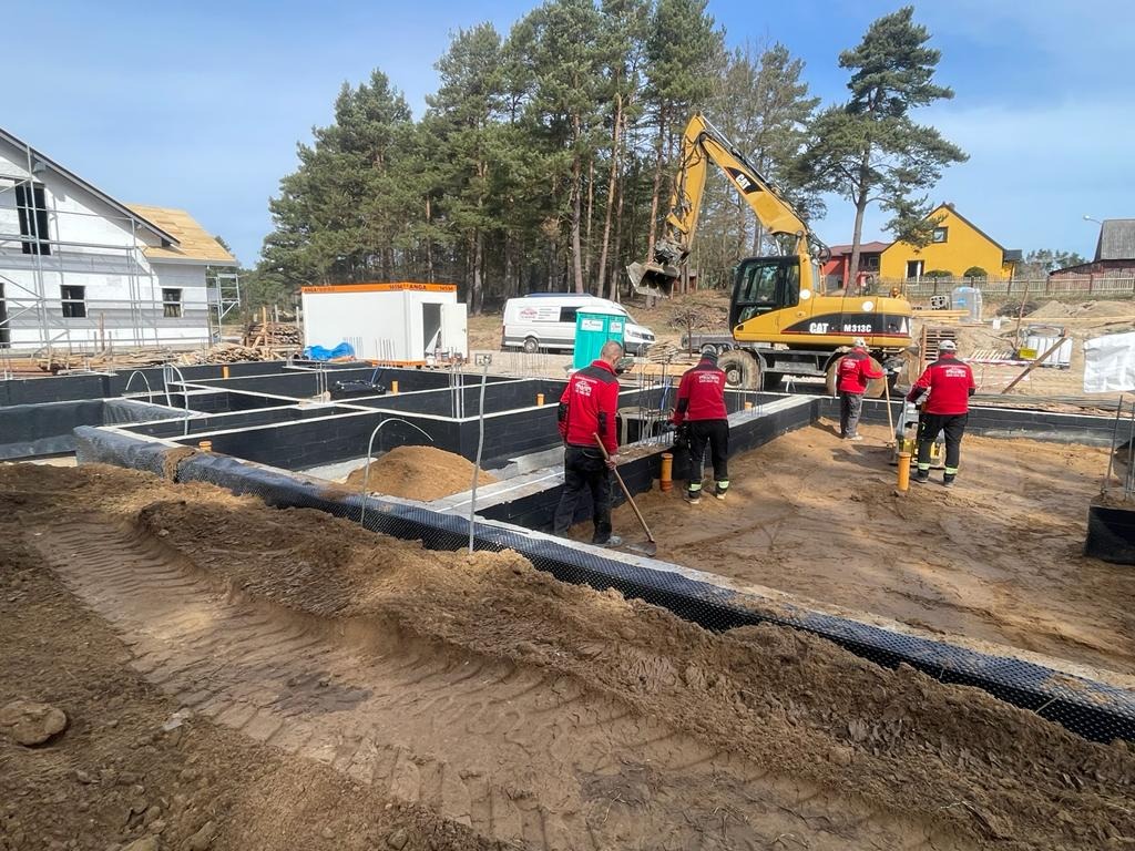 Ekipa budowlana w czerwonych uniformach pracuje przy budowie fundamentów domu jednorodzinnego, widoczny wykop, izolacja przeciwwilgociowa, szalunki, koparka CAT M313C w tle, częściowo zadaszony...