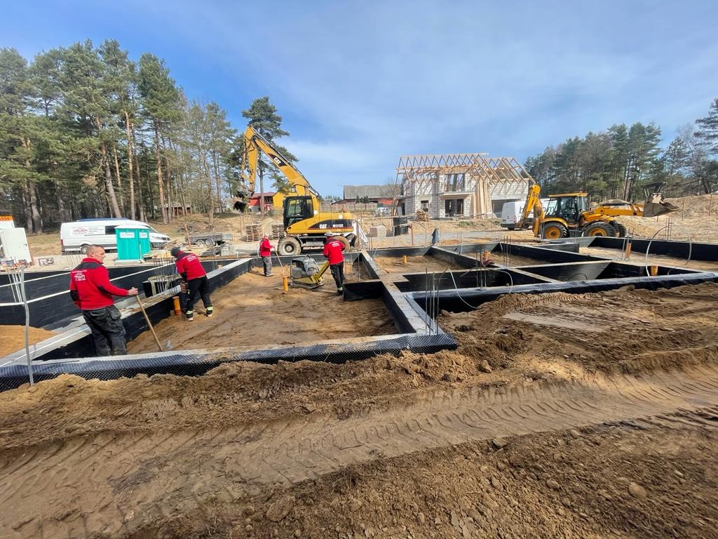 Budowa fundamentów domu z widocznym zbrojeniem, izolacją, pracownikami w czerwonych uniformach, koparką i częściowo wzniesioną konstrukcją szkieletową budynku w tle.