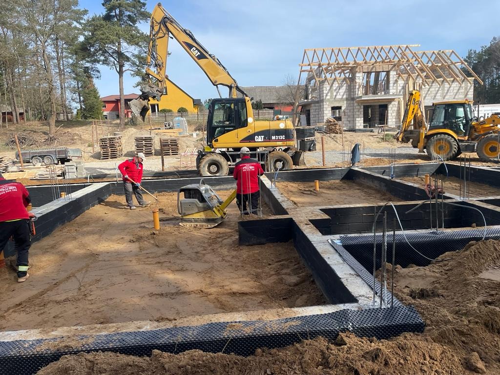 Przygotowanie terenu pod fundamenty domu murowanego: ubijanie piasku zagęszczarką, widoczne zbrojenia, izolacja pionowa fundamentów oraz częściowo wzniesione ściany z pustaków ceramicznych i więźba...