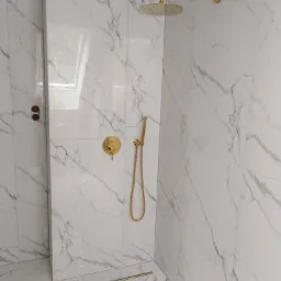 Kabina prysznicowa wykończona białymi płytkami imitującymi marmur, z złotą armaturą: deszczownicą, baterią i odpływem liniowym. W tle widoczne okno dachowe.