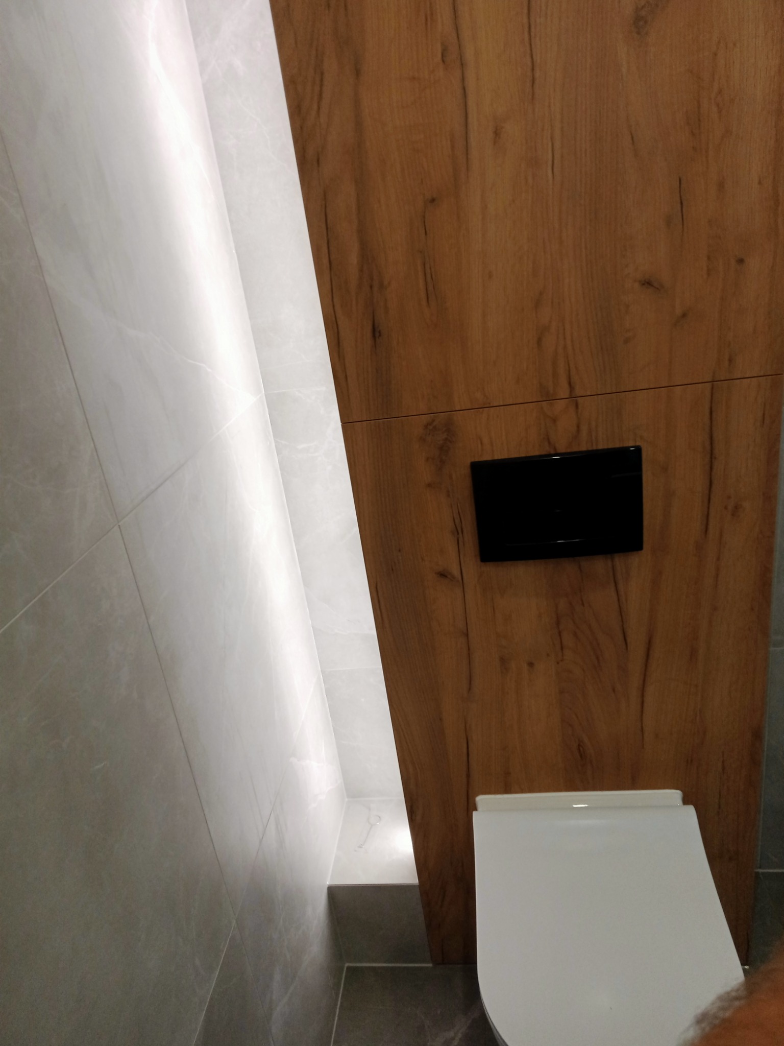 Nowoczesna łazienka z drewnianą zabudową, podświetlaną wnęką i białymi płytkami imitującymi marmur oraz wiszącą miską WC z czarnym przyciskiem.