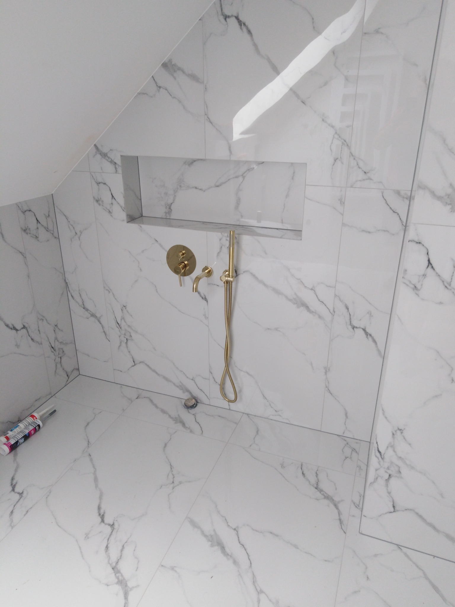 Nowoczesna kabina prysznicowa w poddaszu wykończona białymi płytkami imitującymi marmur, z zamontowaną złotą armaturą i wnęką w ścianie.