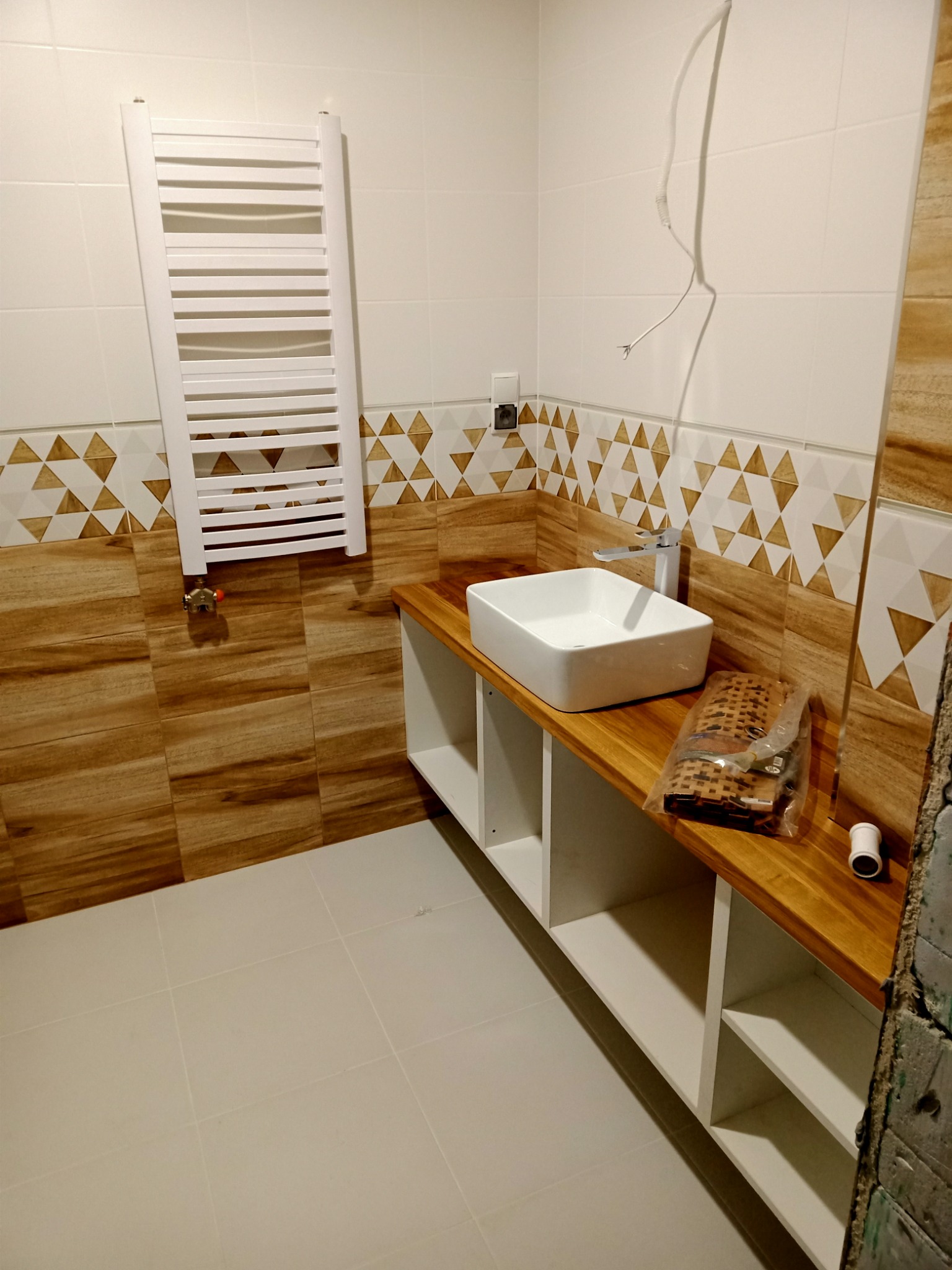 Wnętrze łazienki w trakcie wykańczania, widoczna drewniana umywalka z białą, kwadratową misą, biały grzejnik drabinkowy na ścianie z dekoracyjnymi płytkami, oraz wystające przewody elektryczne.