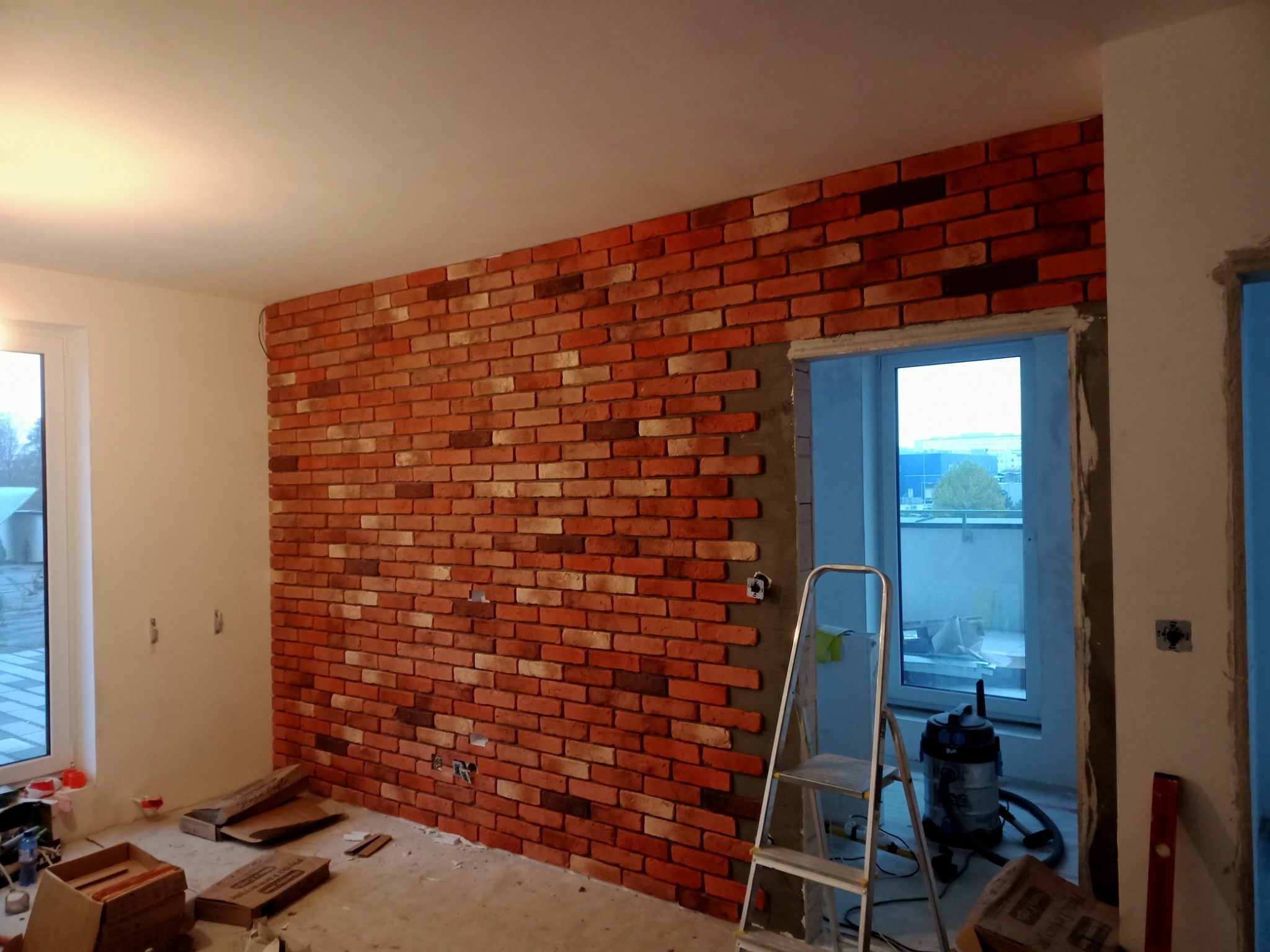 Ściana z cegły klinkierowej w trakcie montażu, widoczne puszki elektryczne, drabina aluminiowa i odkurzacz przemysłowy w pomieszczeniu w stanie deweloperskim.