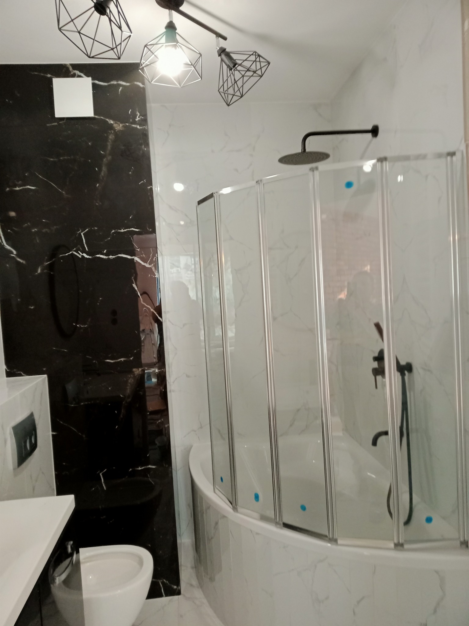 Nowoczesna łazienka z czarno-białymi marmurowymi płytkami, kabiną nawannową ze składanymi drzwiami i geometrycznymi lampami sufitowymi.