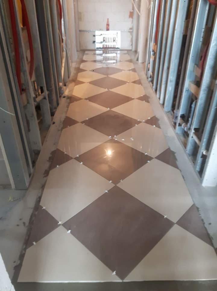 Układanie podłogi z płytek w romby, jasne i brązowe, z użyciem krzyżyków dystansowych, widoczne profile metalowe wzdłuż ścian.