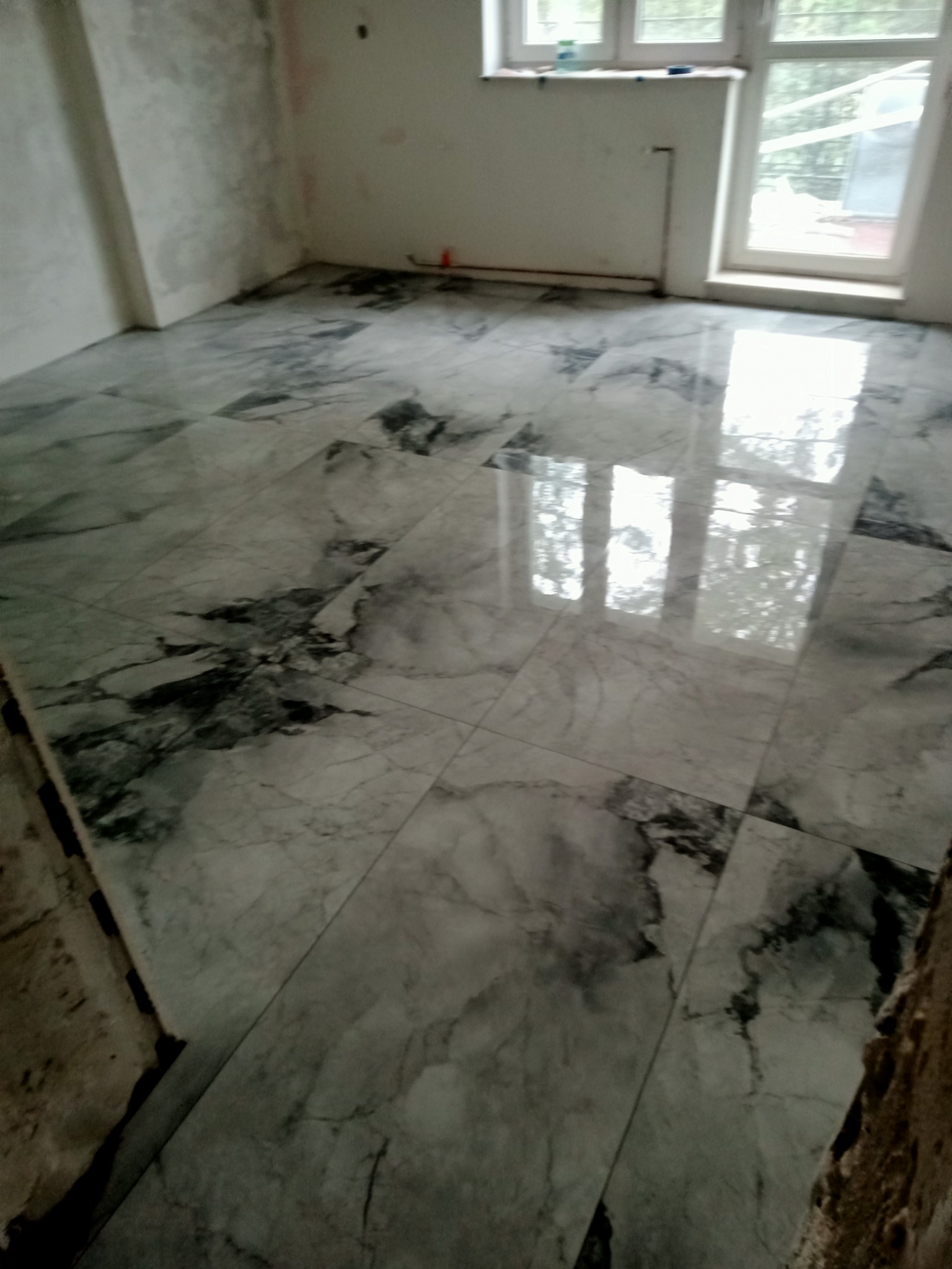 Ułożona podłoga z płytek imitujących marmur w surowym pomieszczeniu z oknem, widoczne niedokończone ściany.