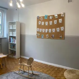 Jasne, przestronne wnętrze sali zabaw z drewnianym koniem na biegunach na szarym dywanie, korkową tablicą z literami i zdjęciami dzieci oraz białym regałem z książkami i zabawkami.