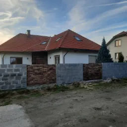 Wznoszony mur z cegły klinkierowej pomiędzy betonowymi słupkami, na tle białego domu z czerwoną dachówką i błękitnego nieba z chmurami.