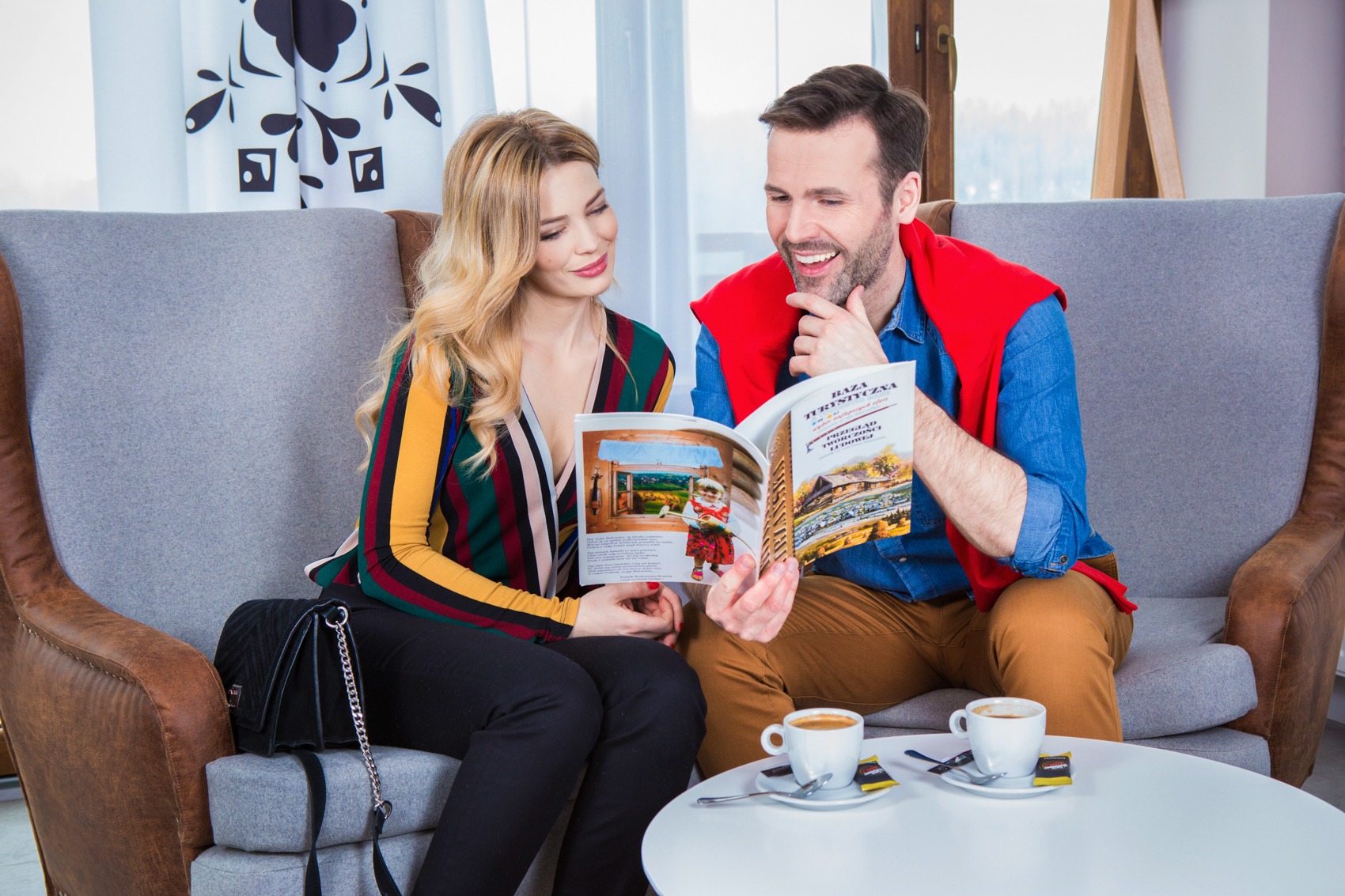 Para siedzi na szarym fotelu i przegląda katalog turystyczny z drewnianą chatą, na stole dwie filiżanki z kawą.
