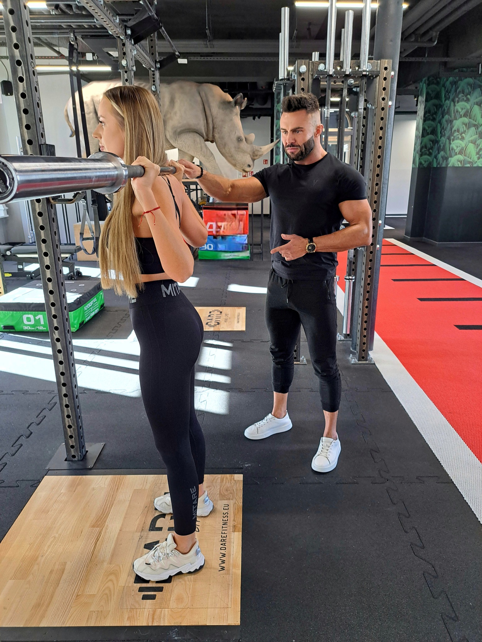 Kobieta ćwiczy przysiady ze sztangą pod okiem trenera w siłowni z rzeźbą nosorożca w tle. Trener wskazuje na pozycję kobiety.