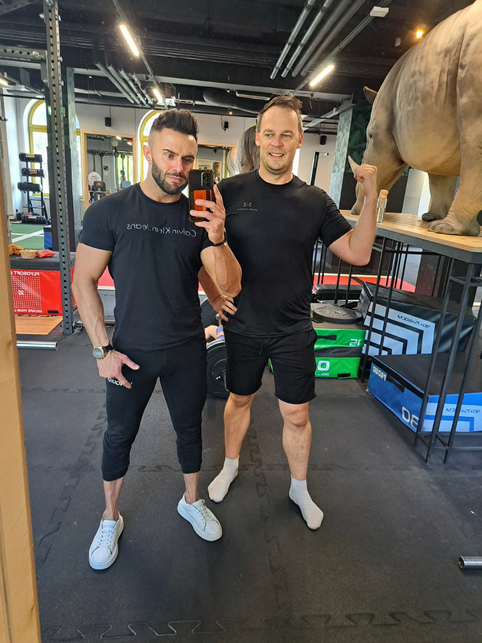 Dwaj mężczyźni w sportowych strojach pozują w siłowni, jeden robi selfie. W tle sprzęt do ćwiczeń i rzeźba nosorożca. Wyjątkowy wystrój siłowni.