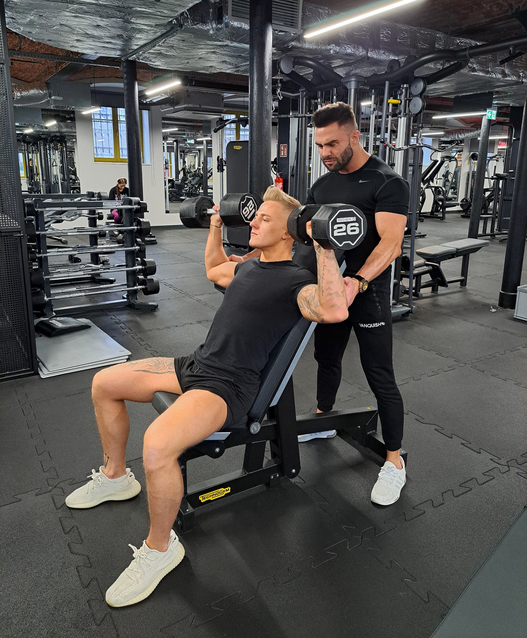 Mężczyzna ćwiczy wyciskanie hantli pod nadzorem trenera w czarnym stroju na siłowni. Widoczne tatuaże, sprzęt do ćwiczeń i okno w tle.