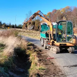 Pomarańczowa minikoparka na gąsienicach ładuje wykopaną ziemię z rowu do zielonej skrzyni ciężarówki na tle błękitnego nieba i jesiennych drzew.