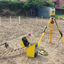 Sprzęt pomiarowy na placu budowy: żółty tachimetr na statywie, tyczka geodezyjna z odbiornikiem GPS i otwarty kufer transportowy. W tle wytyczone punkty.