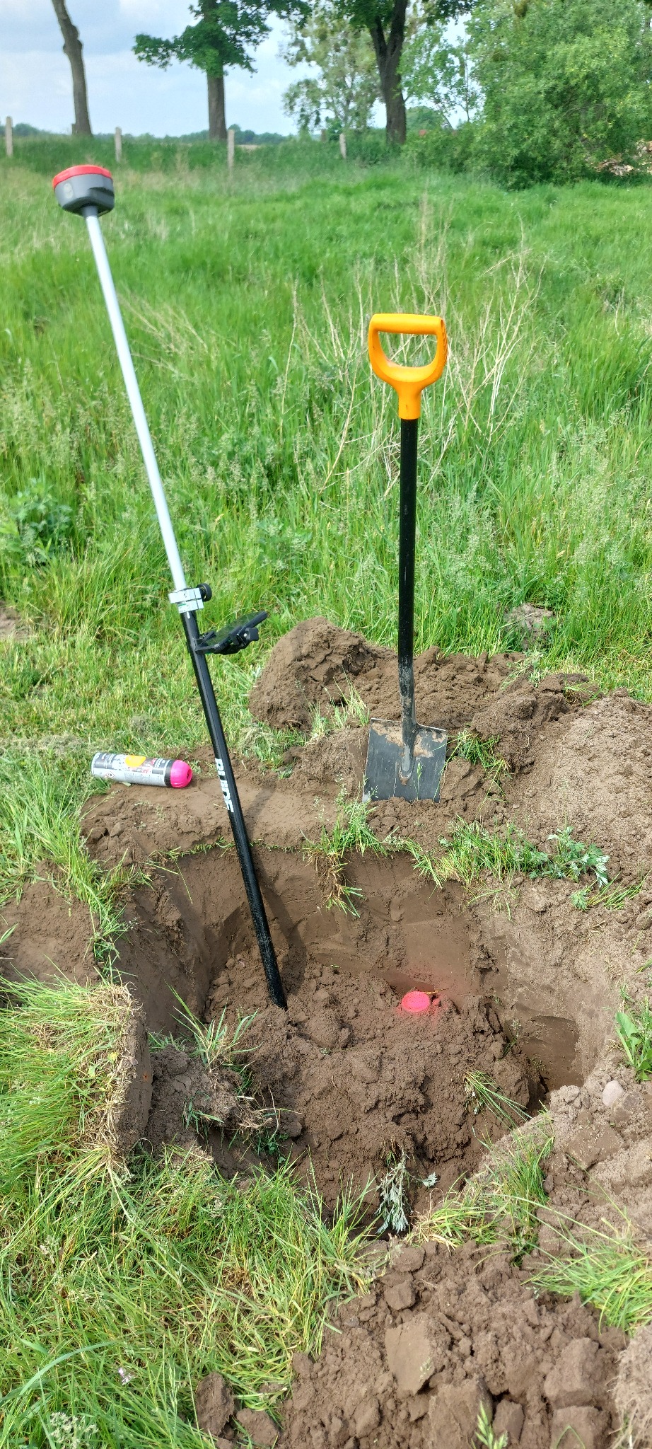 Prace ziemne z użyciem łopaty i tyczki geodezyjnej. W wykopie widoczny pomarańczowy znacznik. Na ziemi leży spray do oznaczania.