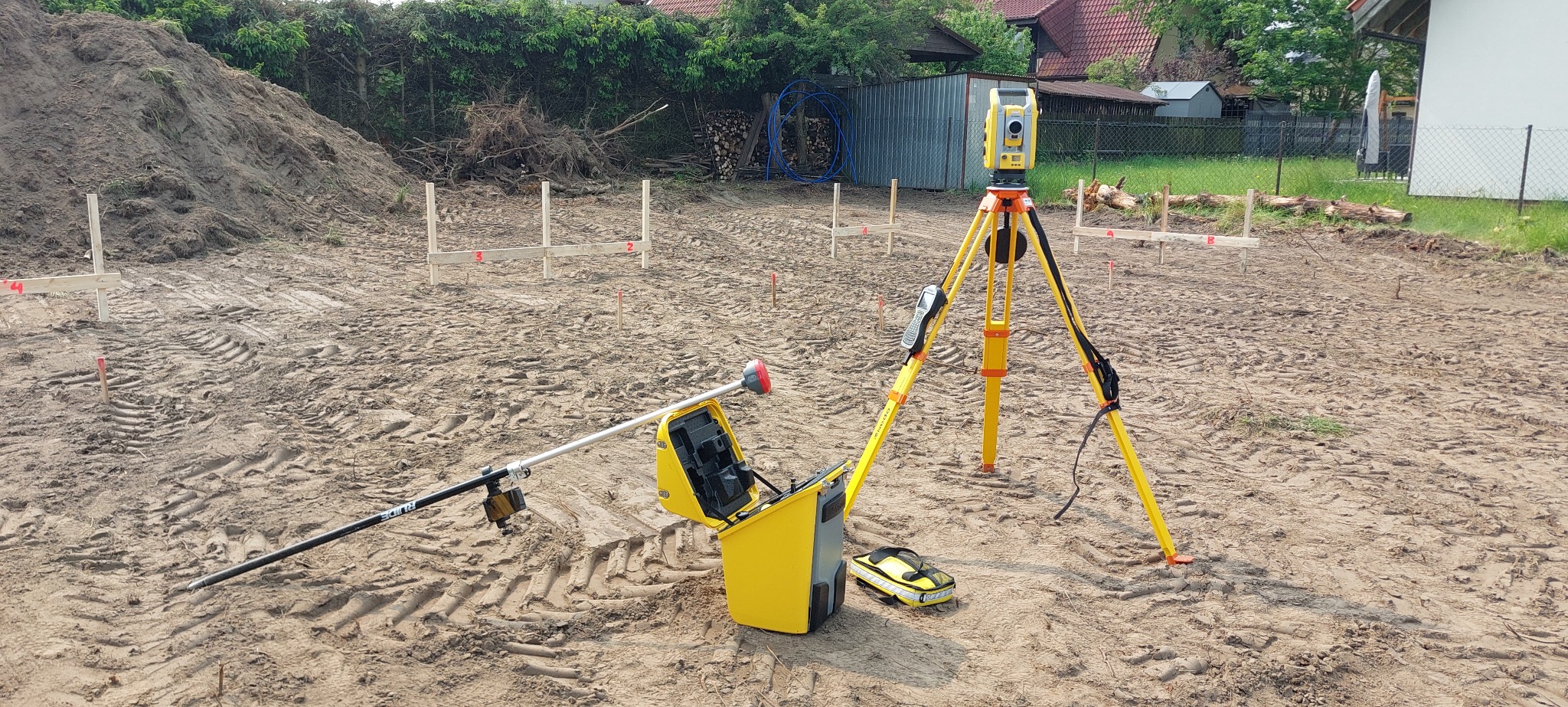 Sprzęt pomiarowy na placu budowy: żółty tachimetr na statywie, tyczka geodezyjna z odbiornikiem GPS i otwarty kufer transportowy. W tle wytyczone punkty.