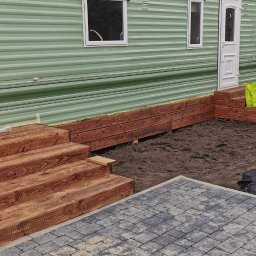 A. Kroczak - Drewniane schody tarasowe w trakcie budowy przy mobilnym domu w Warszawie, z widocznym fragmentem ułożonej kostki brukowej i rozkopanym terenem.