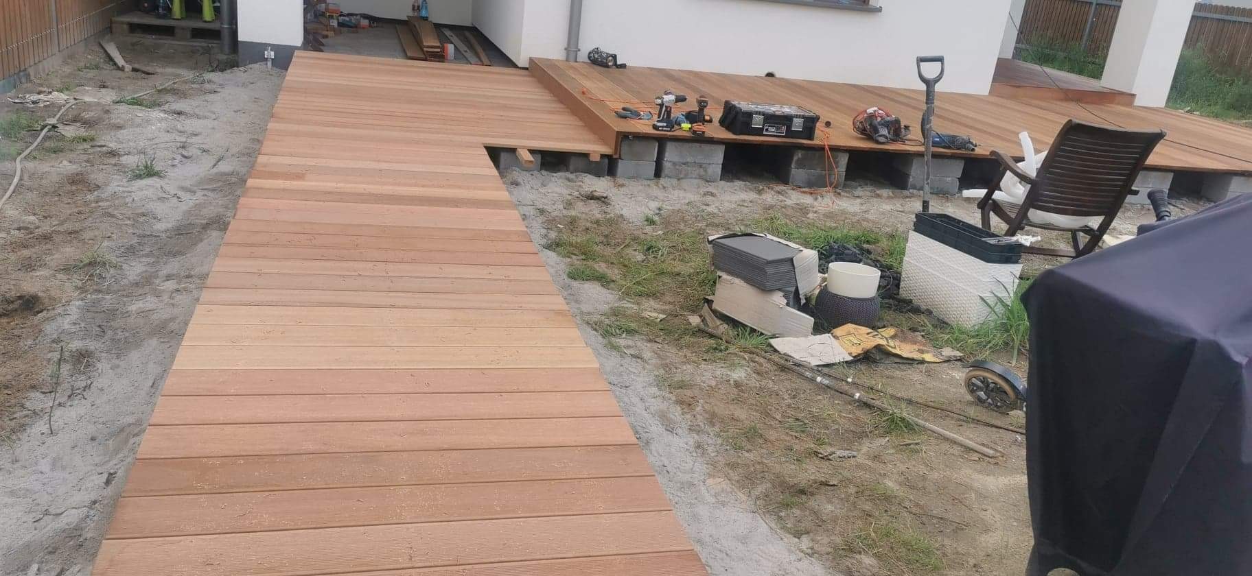 Budowa tarasu z desek drewnianych, widoczne narzędzia, betonowe podpory i fragment ogrodu w trakcie prac.