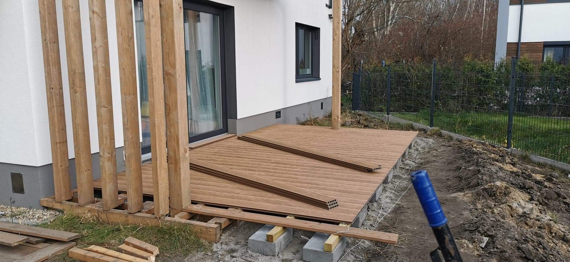 Montaż tarasu kompozytowego przy białym domu z czarnymi oknami, widoczne słupki pergoli i elementy konstrukcji, teren wokół w trakcie prac ziemnych.