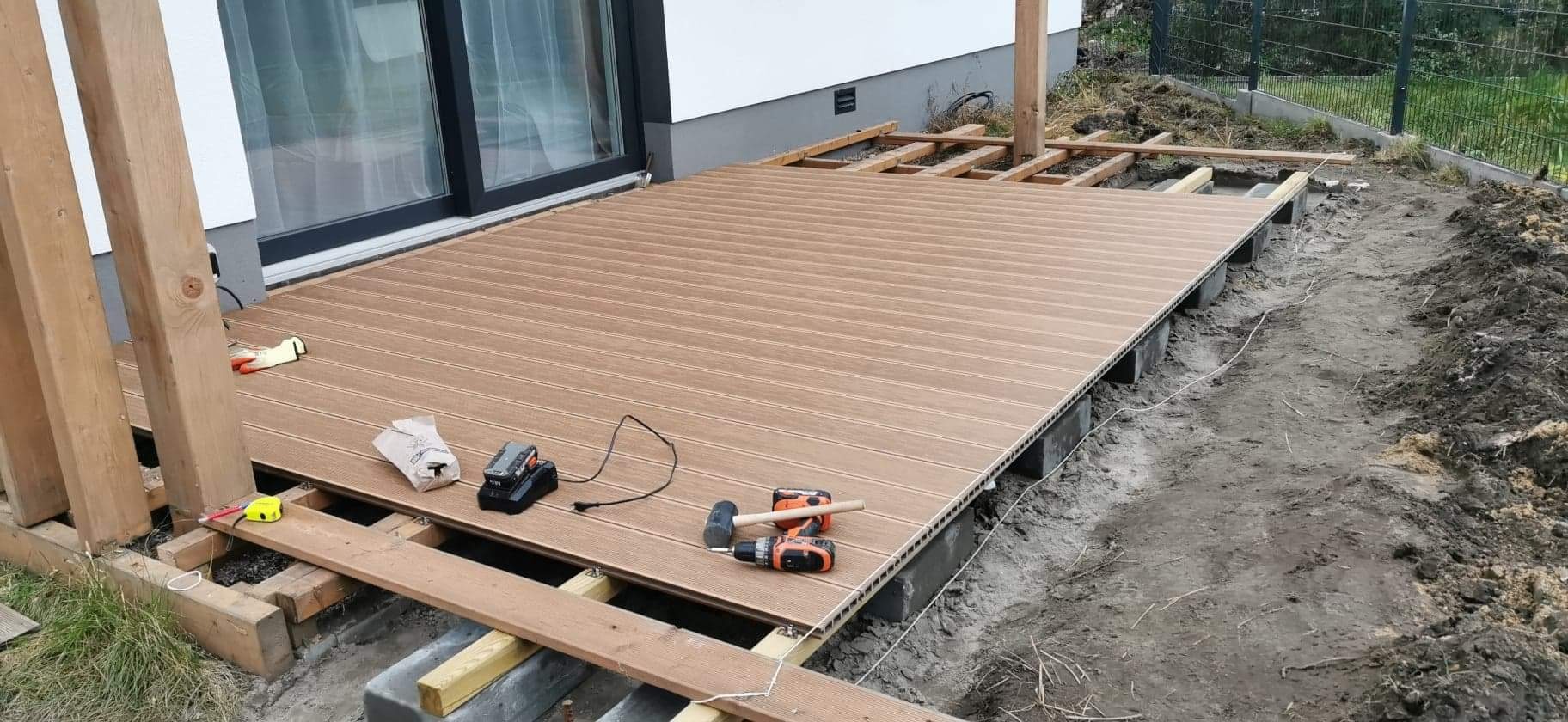 Montaż tarasu kompozytowego: widoczna konstrukcja nośna, częściowo ułożona deska, wkrętarka i młotek obok, teren wokół przygotowany pod dalsze prace.