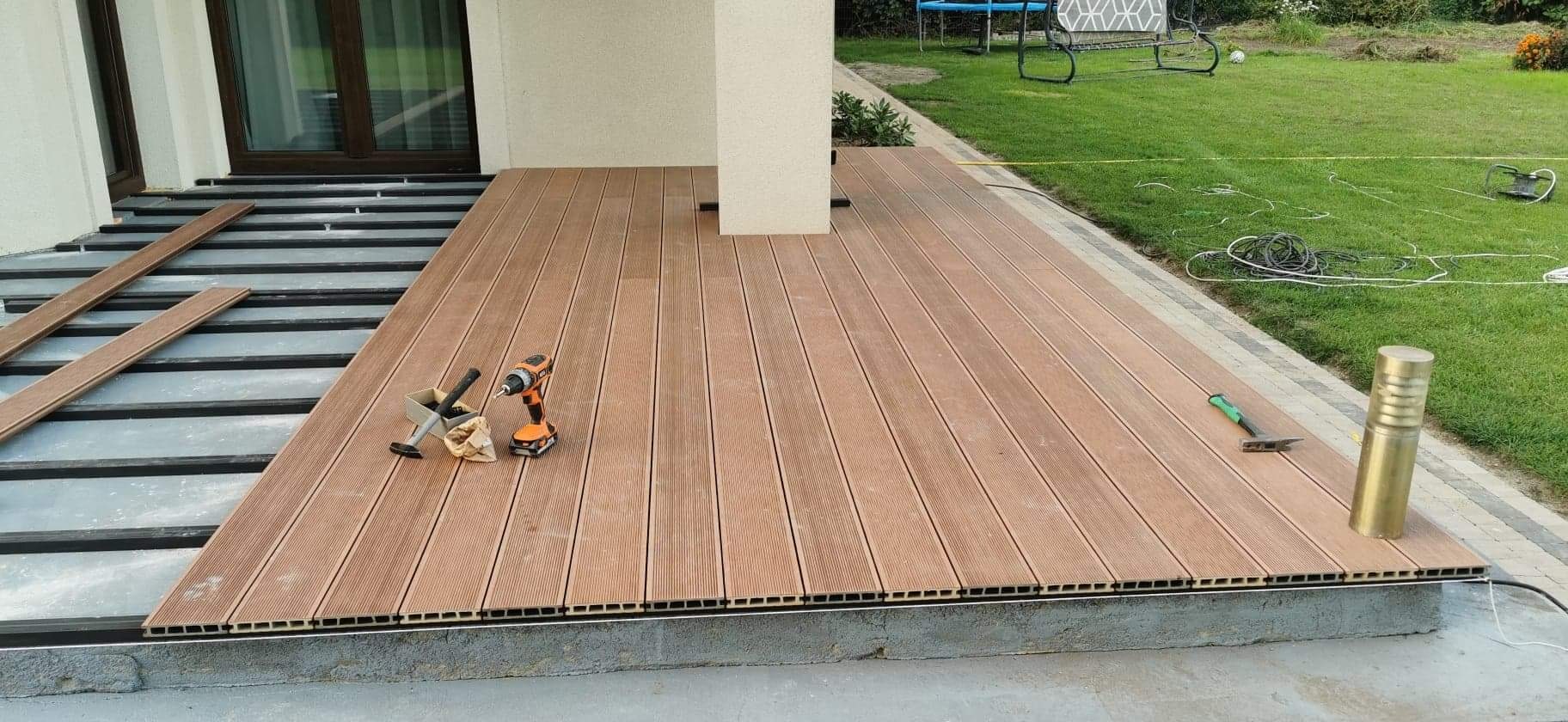 Montaż tarasu kompozytowego: widoczne deski, podkonstrukcja, wkrętarka i narzędzia na częściowo ułożonej powierzchni, z ogrodem w tle.
