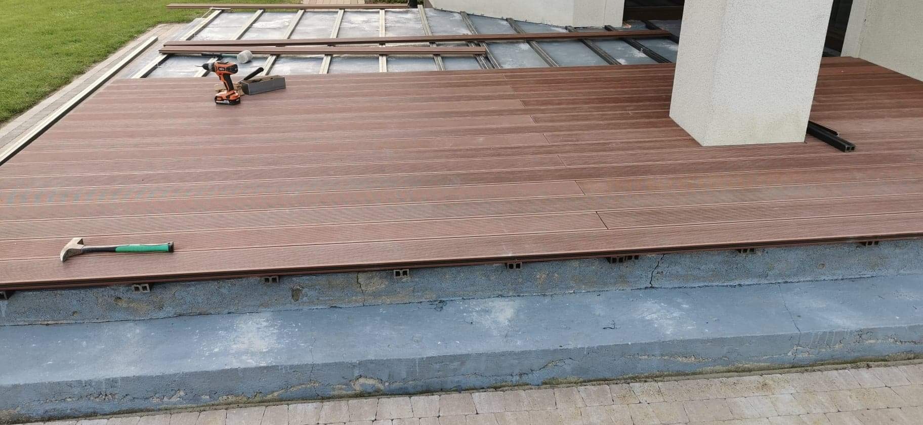 Montaż brązowego tarasu kompozytowego na betonowej wylewce, widoczna wkrętarka akumulatorowa i młotek z zieloną rękojeścią na częściowo ułożonych deskach.