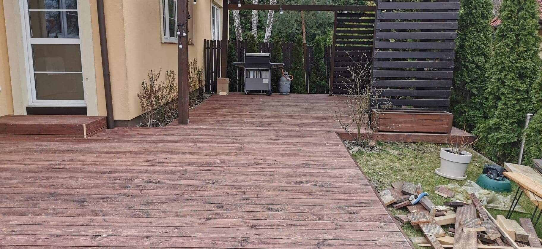 Ciemnobrązowy taras drewniany przylegający do domu z żółtą elewacją, z elementami pergoli, grillem i nieuprzątniętymi resztkami materiałów budowlanych.