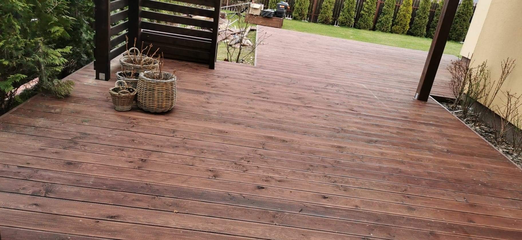 Ciemnobrązowy taras z desek drewnianych, z trzema wiklinowymi koszykami na rośliny w rogu, widok na trawnik i tuje w tle.