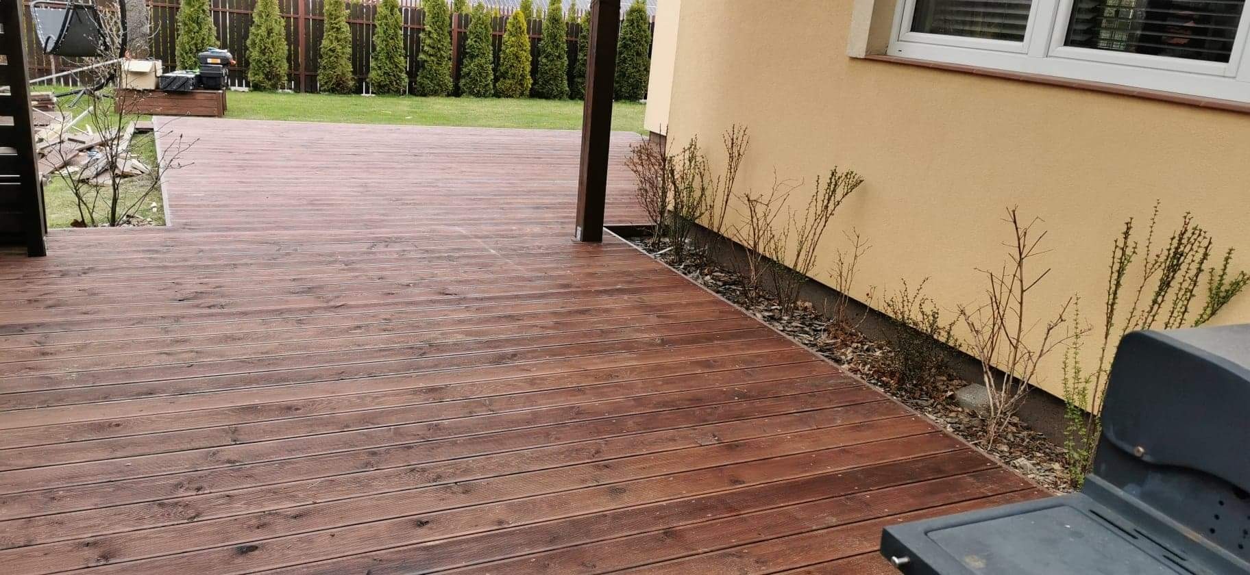 Ciemnobrązowy taras drewniany z desek ryflowanych, przylegający do ściany budynku z niską rabatą z krzewami i korą, w tle żywopłot z tuj.