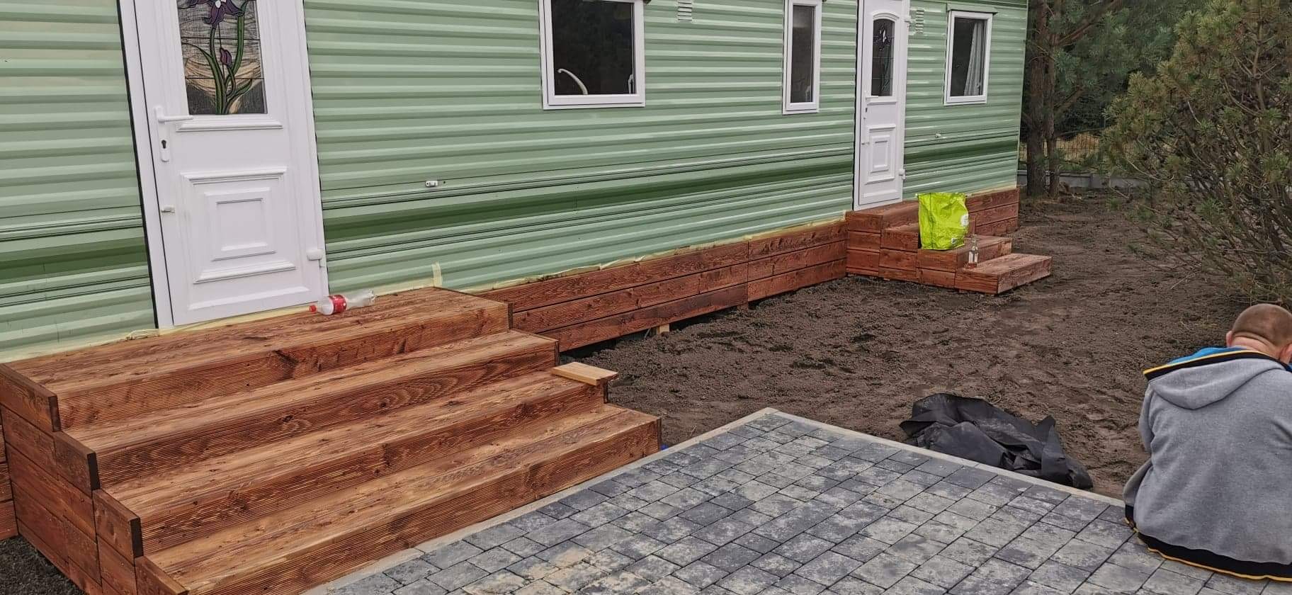 Drewniane schody tarasowe w trakcie budowy przy mobilnym domu w Warszawie, z widocznym fragmentem ułożonej kostki brukowej i rozkopanym terenem.