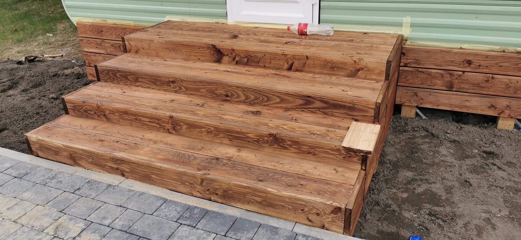 Schody z drewna prowadzące do drzwi, z butelką plastikową na górnym stopniu, widoczne niedociągnięcia w wykończeniu i świeża ziemia wokół.