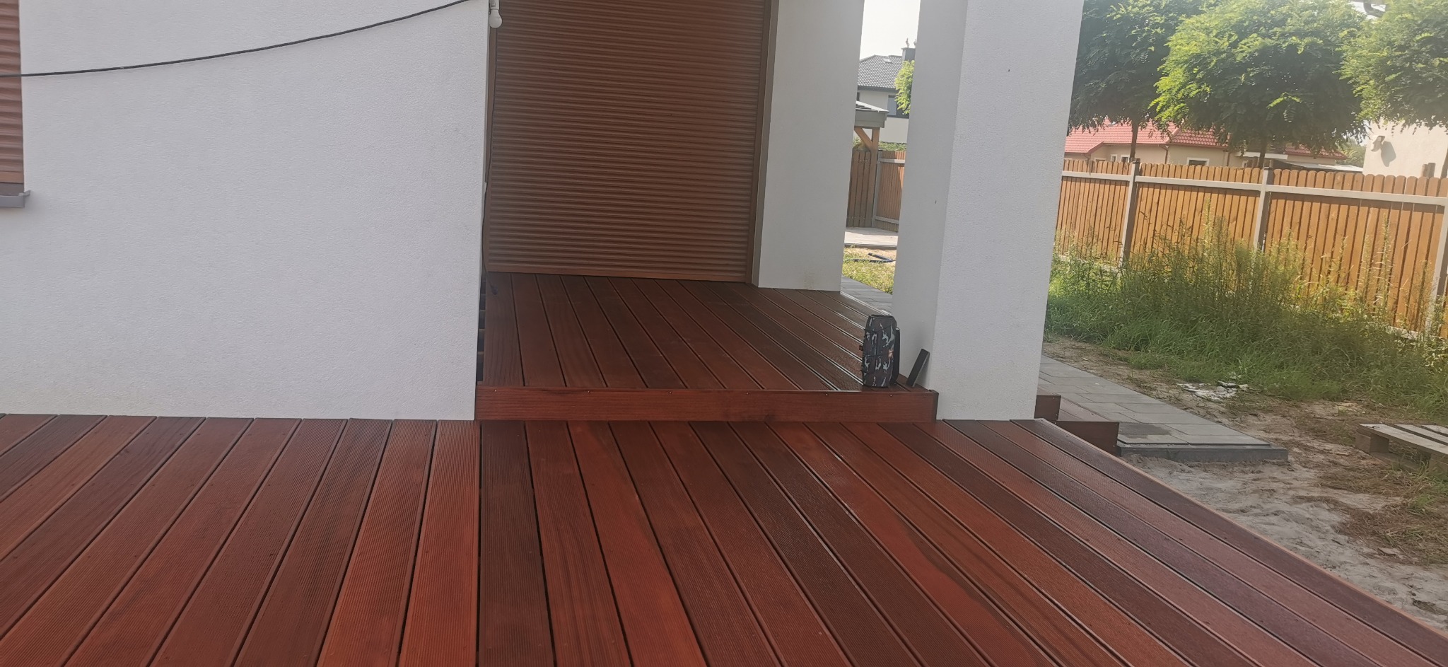 Ciemnobrązowy taras z desek, z widoczną strukturą drewna, przylegający do białego budynku z roletami, z widocznym fragmentem ogrodu w tle.