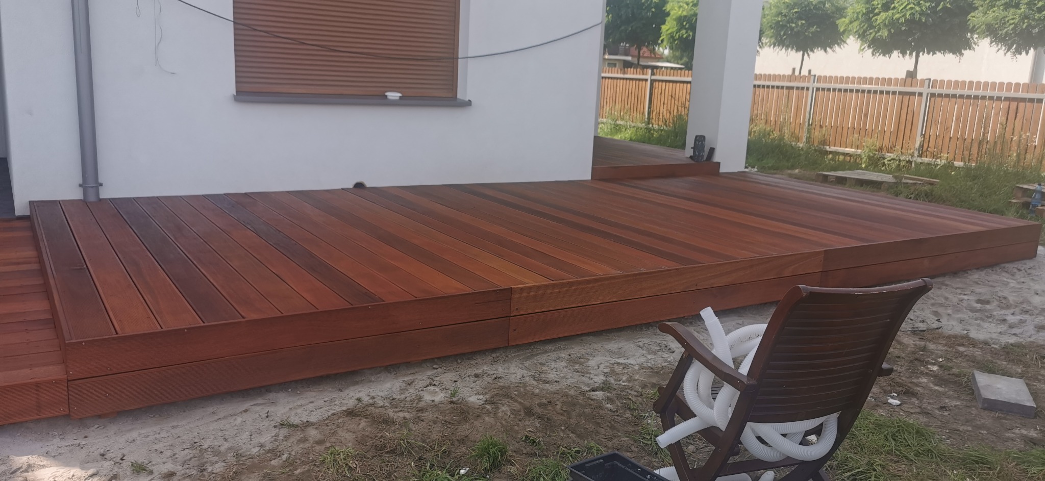 Nowoczesny taras z drewna egzotycznego, z widocznymi poziomami i schodkami, częściowo ukończony, z elementami wykończeniowymi w postaci krzesła i węża ogrodowego.