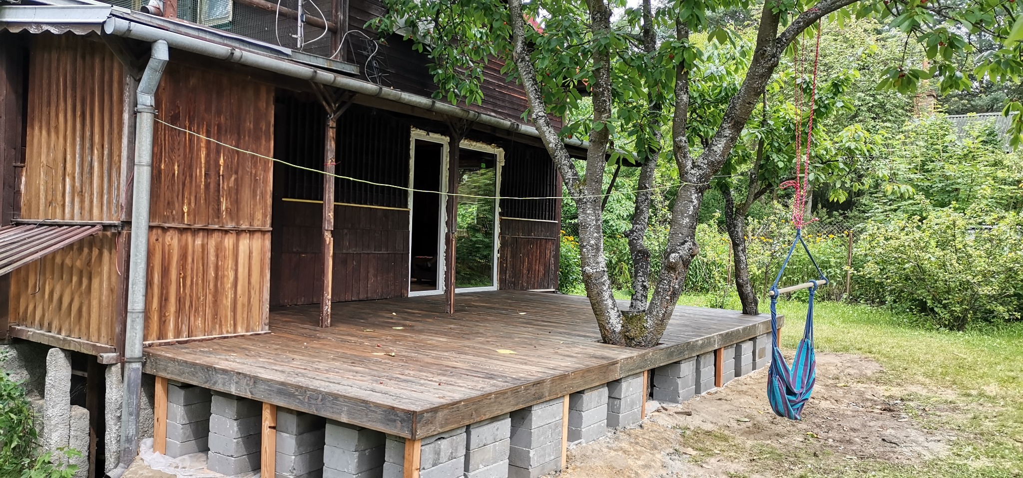 Drewniany taras z drzewami rosnącymi przez deski, wsparty na betonowych blokach, z huśtawką hamakową wiszącą obok, w tle drewniany dom z białymi ramami okiennymi i bujna zieleń ogrodu.