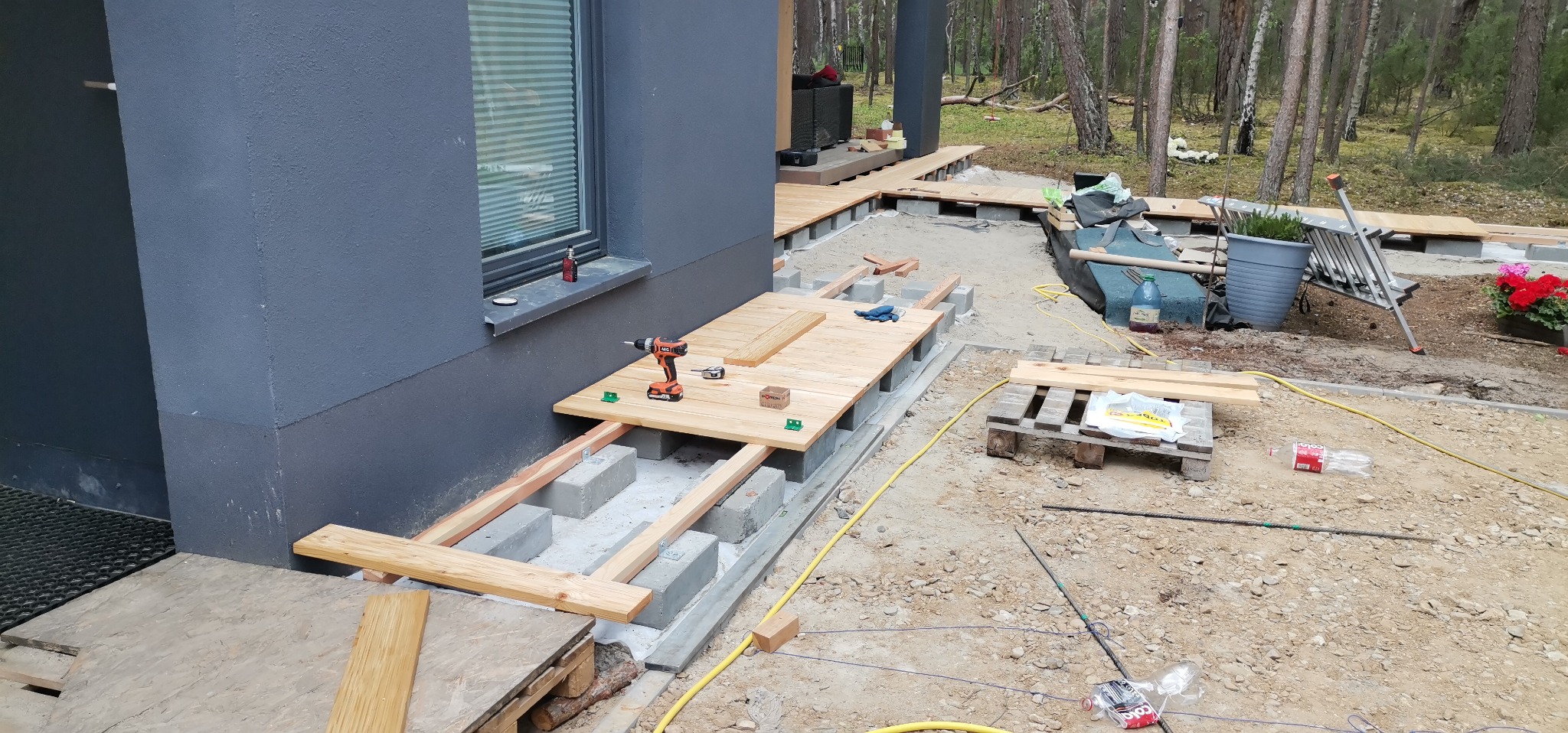 Budowa tarasu drewnianego przy szarym domu z widocznymi betonowymi bloczkami, drewnianą konstrukcją nośną, deskami, wkrętarką i zielonymi podkładkami dystansowymi na tle leśnym.
